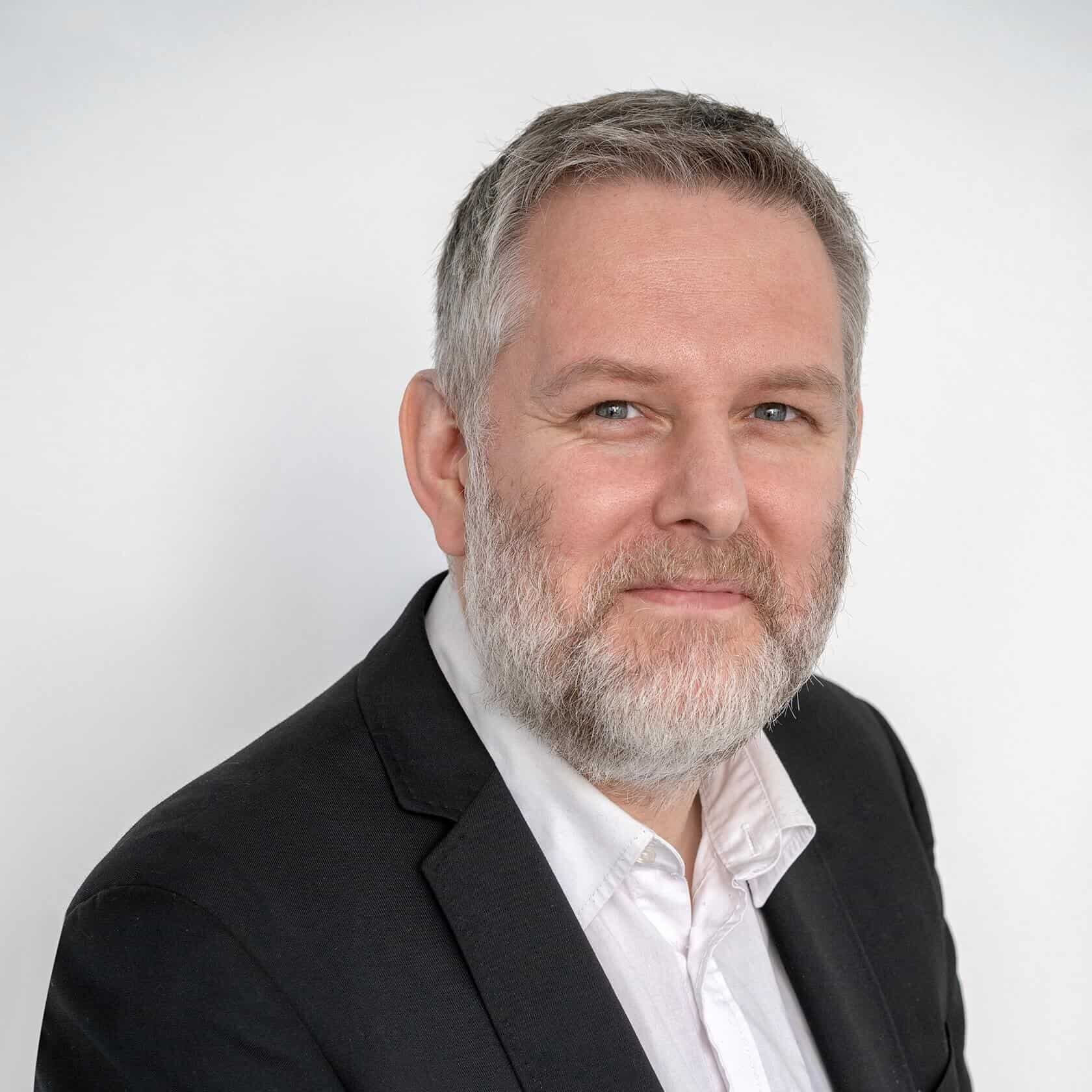 A person wearing a dark suit jacket and a light collared shirt is shown from the chest upward against a plain background.
