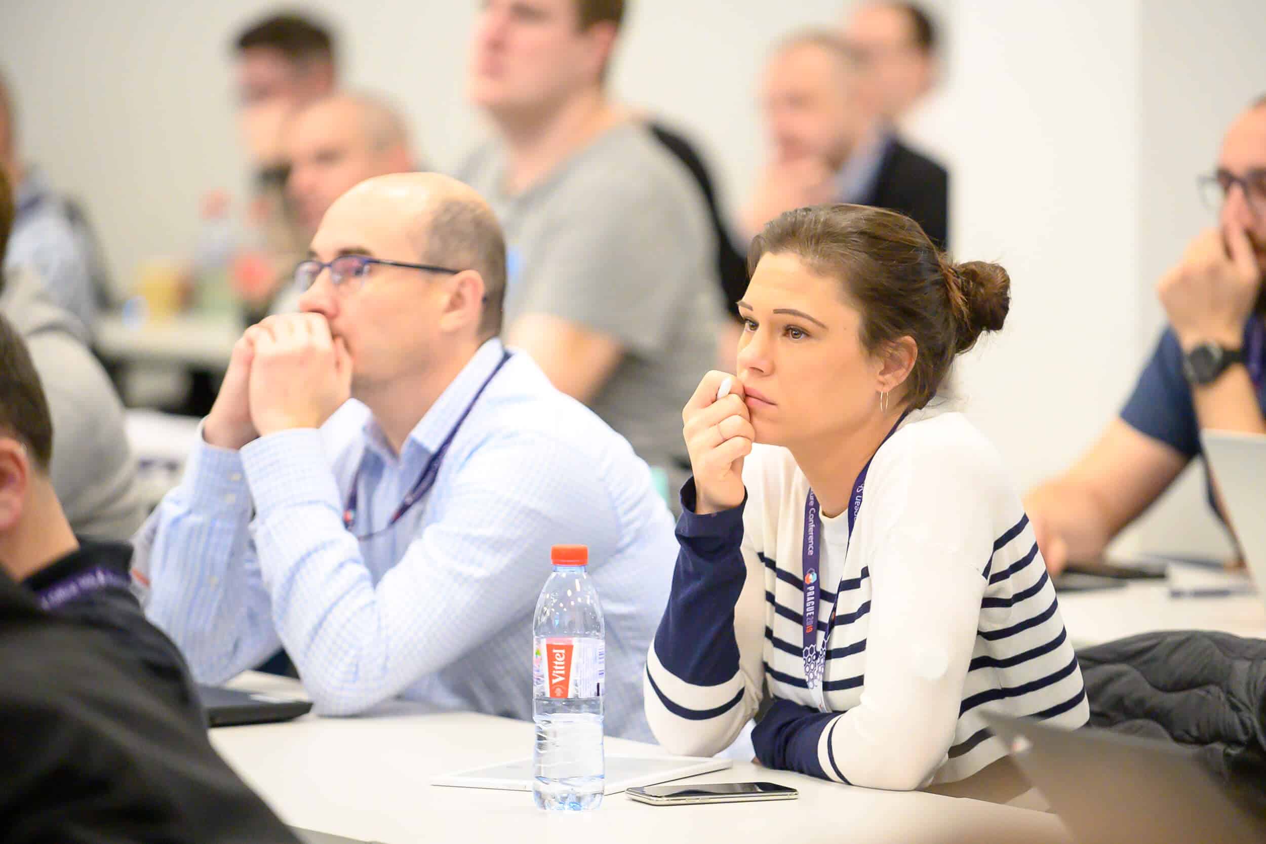Several people sit at tables in a classroom or conference setting, facing toward the front of the room. Some individuals have laptops open, and a water bottle is visible on one of the desks. The environment appears bright and focused, with participants seated in rows