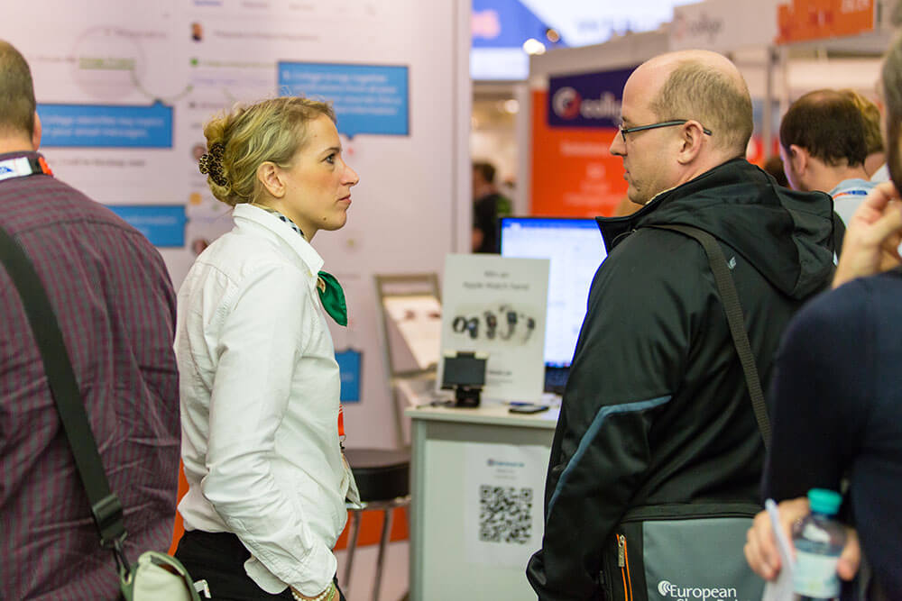 Two people stand and talk in an exhibition or conference space. A booth with informational posters, a display screen, and a counter featuring promotional materials is visible behind them. Additional attendees and displays can be seen throughout the background.