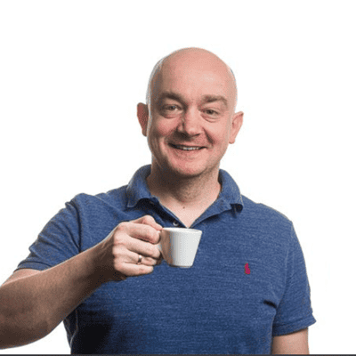 A person wearing a short‑sleeved polo shirt is standing against a plain white background and holding a small cup in one hand, raised near the chest.