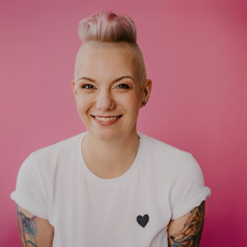 A person wearing a white T‑shirt with a small heart emblem on the chest, seated against a solid pink background