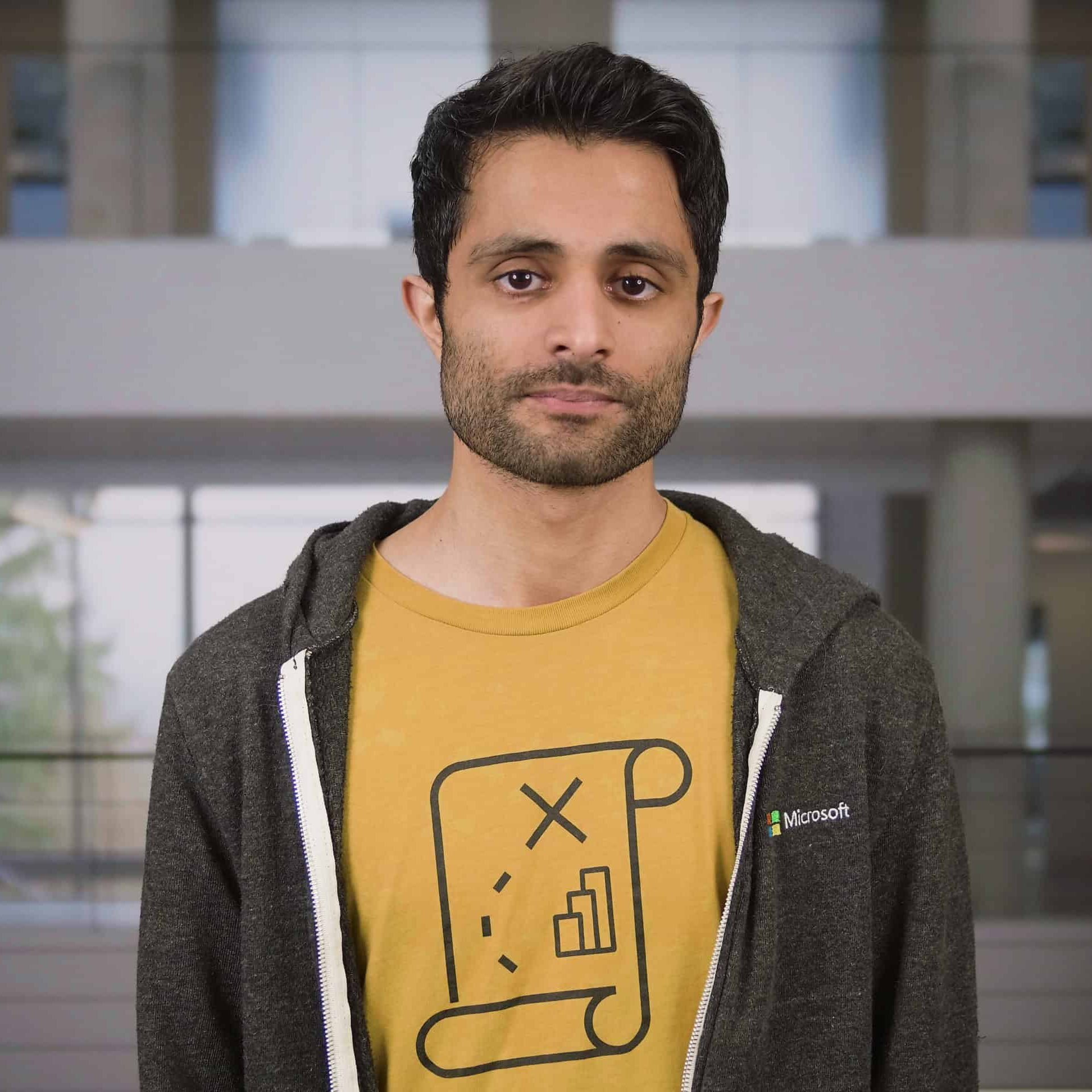 A person is standing indoors wearing a yellow shirt with a printed graphic design and a dark zip‑up hoodie. The background shows a modern building interior with large windows and structural columns.