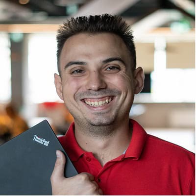 A smiling person with dark hair wearing a red shirt holding a laptop.