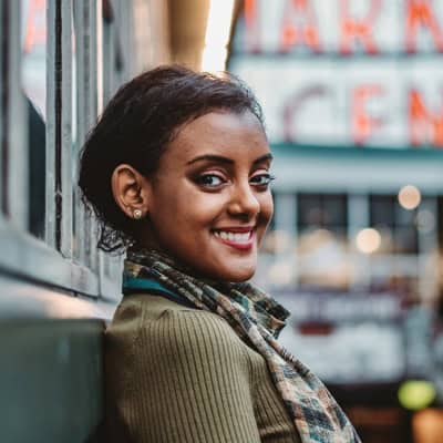 A smiling person with dark hair wearing a green top and checked scarf with city buildings in the background.