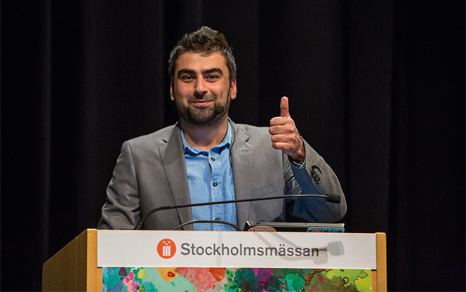speaker looking at the camera and giving a thumbs up while standing at a lectern