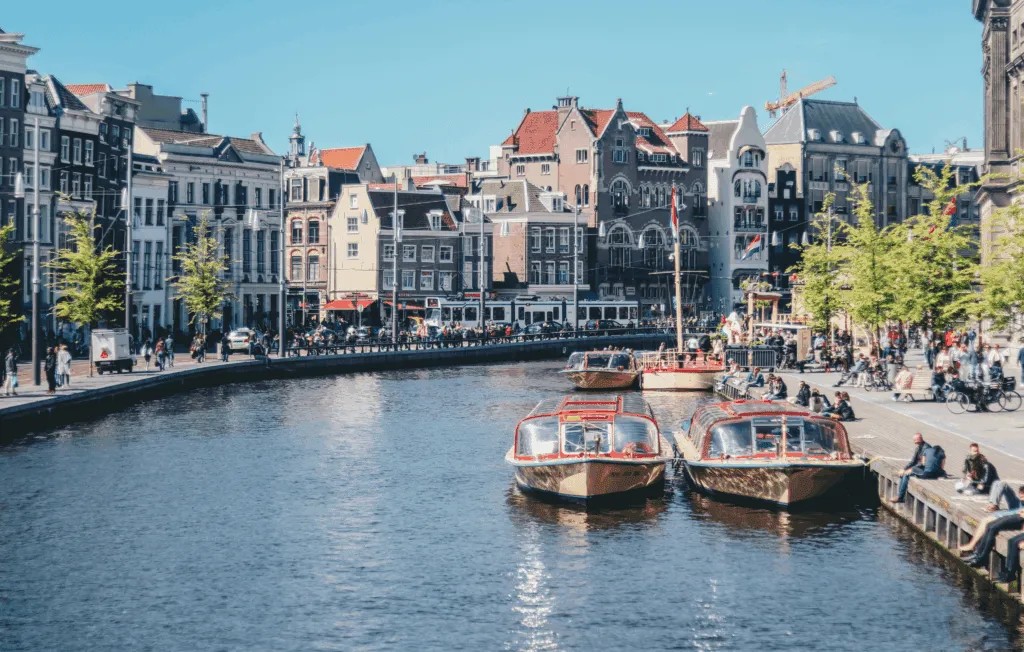 Row of tall, narrow canal houses with large windows along a waterfront in Amsterdam, with trees lining the street, parked bicycles and cars, and several small boats floating on the canal in the foreground.