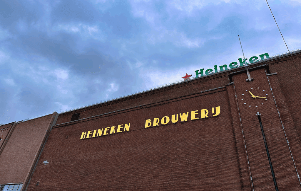 Exterior of a large brick building with yellow letters spelling ‘Heineken Brouwerij’ and a clock on the wall, with a Heineken sign on the roof against a cloudy sky