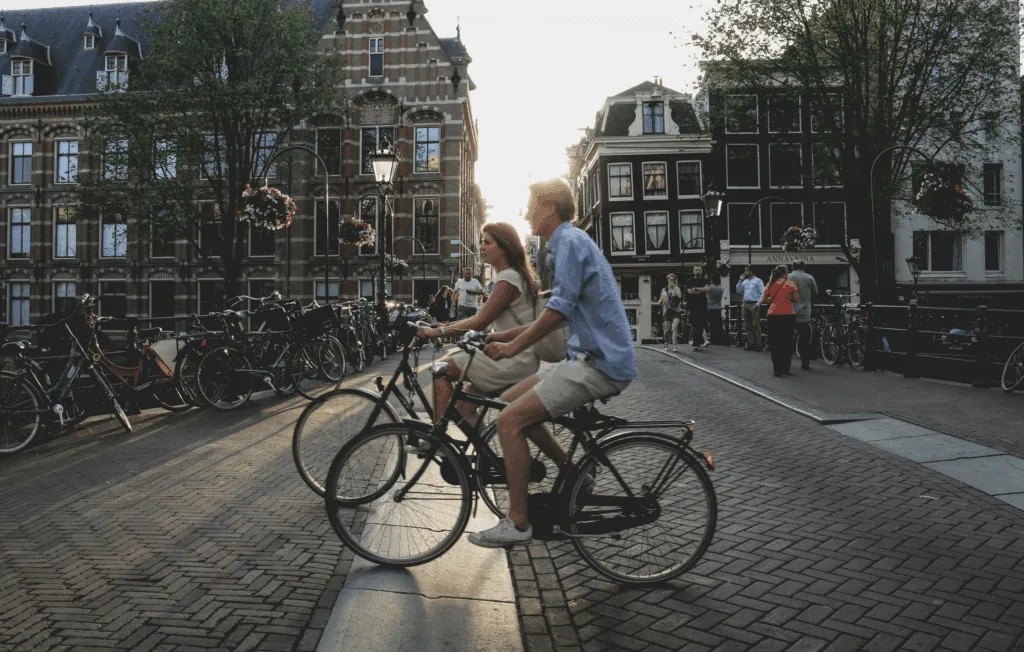 Two people riding bicycles on a cobblestone street in a historic city area lined with tall buildings, with many parked bicycles in the background and the sun shining through the buildings.