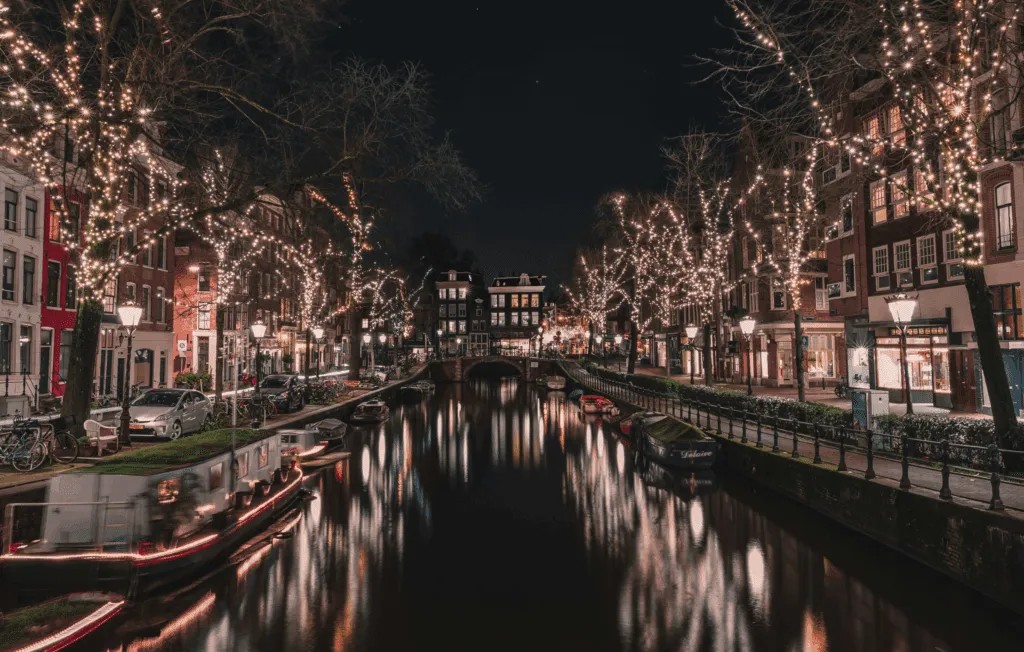 Night‑time view of an Amsterdam canal with reflections of string lights wrapped around trees, historic buildings on both sides, and several moored boats
