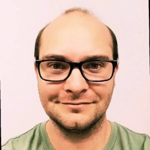 Head‑and‑shoulders portrait of a man wearing black rectangular glasses and a green T‑shirt, facing the camera with a slight smile against a light, plain background.