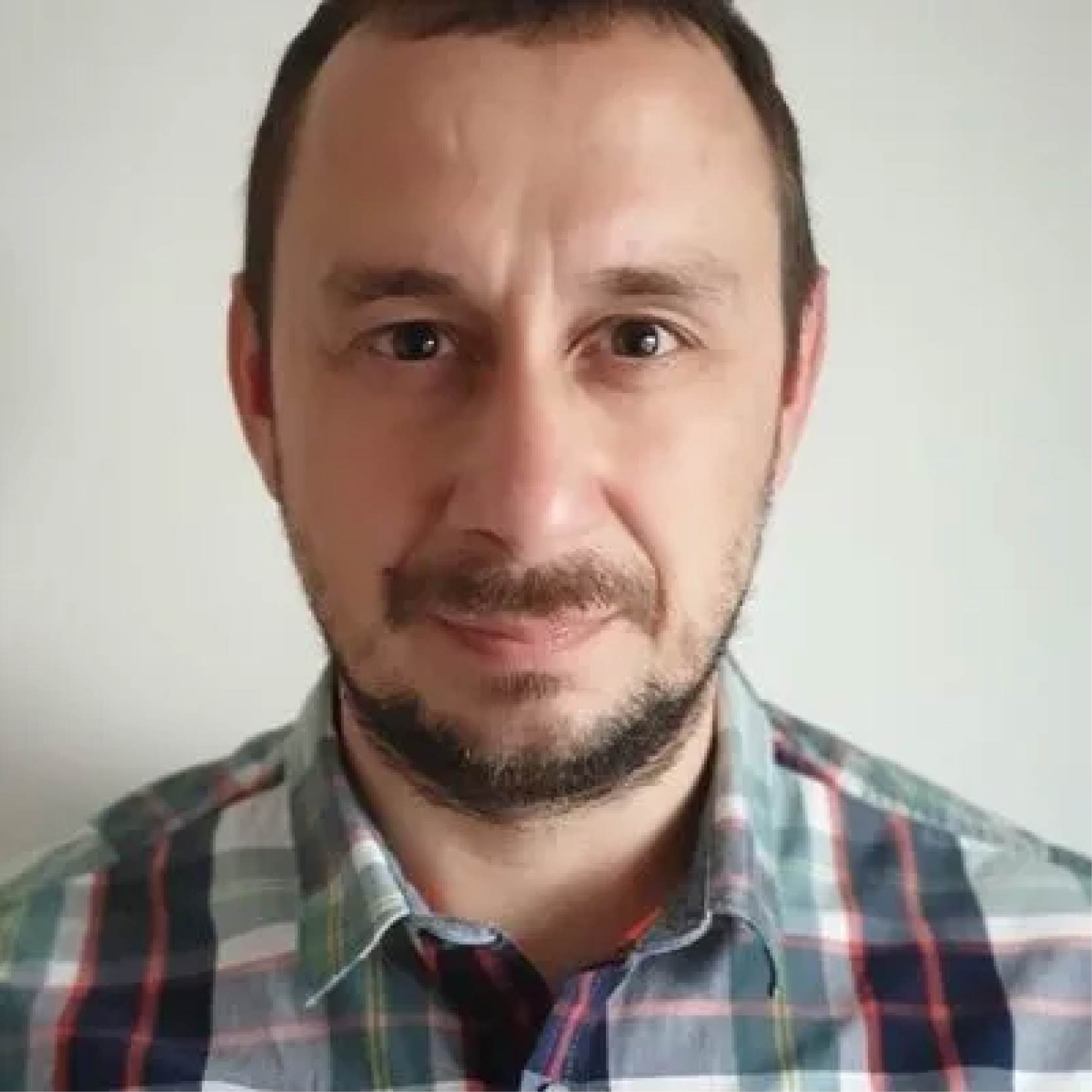 A person is shown from the shoulders up wearing a checkered shirt in shades of green, blue, and red, standing in front of a plain light‑colored wall.”
