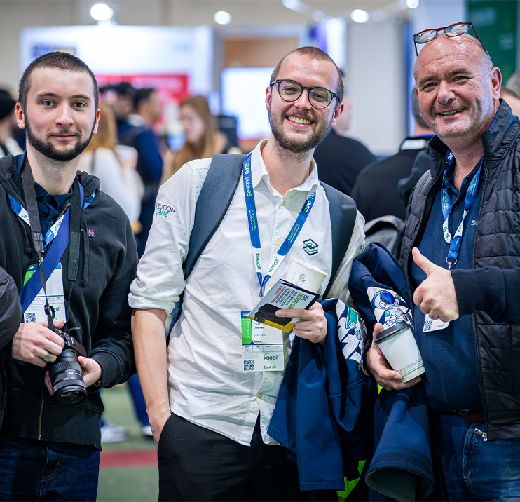 Three attendees pose for the camera while smiling and giving a 'thumbs up' sign, in the EXPO hall
