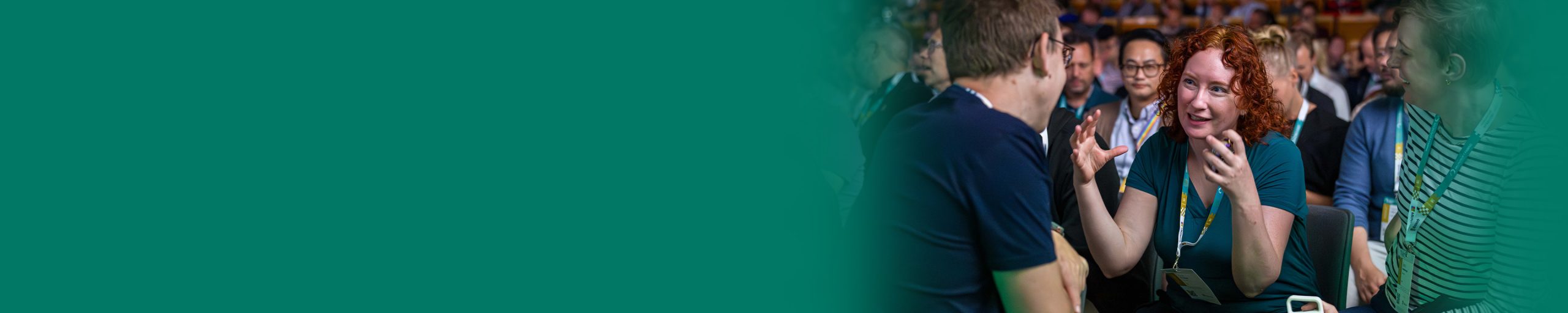 A group of people are gathered in a busy indoor setting. One person in the foreground is holding a phone and appears to be speaking to another person facing them. Several others stand close by, and the background is filled with people and warm lighting.