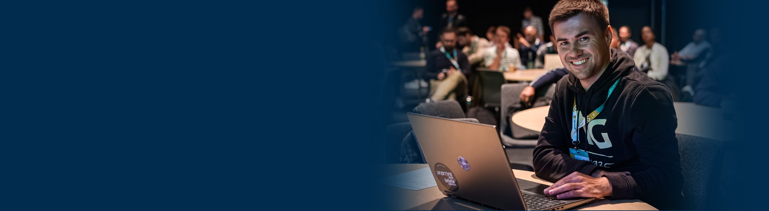 A person sits at a round table using a laptop during an indoor event, with several other attendees seated in the background. The person is wearing a dark hoodie with a lanyard, and the laptop has visible stickers on the back. The scene is set in a large conference-style room with low lighting.